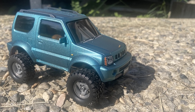 The Jimny is outdoors, driving on a concrete pad. A grass seed has landed on the hood.